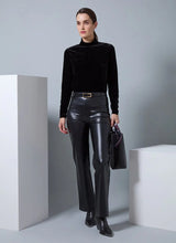 Load image into Gallery viewer, Woman wearing a black velvet top and black leather pants with belt and black boots holding a black handbag against a gray background.