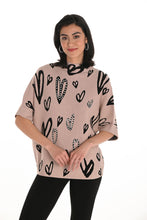 Load image into Gallery viewer, Woman wearing a beige sweater with black heart patterns and rhinestone embellisments, black leggings and black hoop earrings against a white wall.