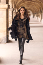 Load image into Gallery viewer, A woman walking through a stone arcade wearing a black faux-fur poncho with pom-poms, a black and gold patterned top, black leather leggings, and black ankle boots.