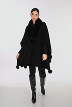 Load image into Gallery viewer, A woman modeling a black poncho with a plush faux-fur collar and pom-pom trim, paired with black leggings and knee-high boots, against a light gray studio background.