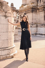 Load image into Gallery viewer, A woman wearing a black sleeveless midi dress with a wide black belt and black knee-high boots, posing outdoors next to a stone column in front of a historic building.