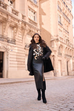Load image into Gallery viewer, A woman stands on a cobblestone street in front of a beige stone building, wearing a black coat, black leather skirt, statement gold and black belt and a black-and-white patterned top with black ankle boots.