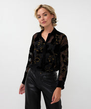 Load image into Gallery viewer, Woman wearing a sheer black blouse with black velour floral pattern and gold lurex threading, black pants and black star earrings against a white background.
