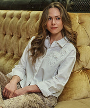 Load image into Gallery viewer, Woman sitting on a plush, gold sofa wearing a white blouse with floral lace details and gold crushed velvet pants.