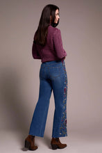 Load image into Gallery viewer, Woman sitting on a stool wearing a purple sweater, blue jeans with floral embroidery and black boots against a neutral background.