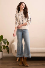 Load image into Gallery viewer, Woman wearing a cream sweater with black crochet stitching throughout, light blue jeans, and brown boots standing in a room with a beige sofa and plant.