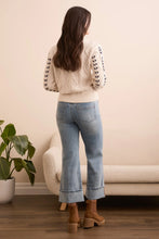 Load image into Gallery viewer, Woman wearing a cream sweater with black crochet stitching throughout, light blue jeans, and brown boots standing in a room with a beige sofa and plant.