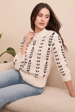 Load image into Gallery viewer, Woman wearing a cream sweater with black crochet stitching throughout and blue jeans sitting on a beige couch.