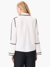 Load image into Gallery viewer, This stunning and unique blouse is a bold choice for fashion lovers who like to stand out. The black trim along the seams creates a striking contrast with the off-white fabric. The pearl buttons add a touch of elegance, while the flounce sleeves have slits at the end for extra flair. The blouse has a classic shirt collar and a hip-length hem.