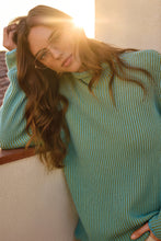 Load image into Gallery viewer, Woman wearing a teal blue sweater and eyeglasses leaning on a ledge with sunlight in the background.