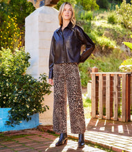Load image into Gallery viewer, A woman stands outdoors wearing a black leather-look button-up shirt and wide-leg cheetah print pants with black ankle boots. The setting includes a stone pillar, greenery, and a wooden fence.