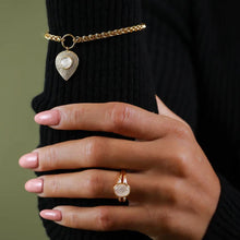 Load image into Gallery viewer, A close-up of a person's hand wearing a gold ring with a round white stone, while the same jewelry set is also shown on the wrist as a gold chain bracelet with a teardrop-shaped pendant featuring a white pearl. The person is wearing a black ribbed sweater against a muted olive green background.