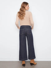 Load image into Gallery viewer, A woman shown from the back wearing dark blue wide-leg cropped jeans, a beige ribbed knit top, and brown mule heels against a plain white wall.
