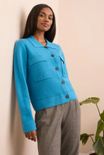 Load image into Gallery viewer, Woman wearing a blue jacket and gray pants standing against a white wall with a plant in the background.