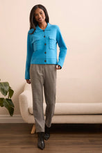Load image into Gallery viewer, Woman wearing a blue jacket, gray pants and black boots standing in a room with a beige sofa and plant.