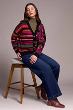 Load image into Gallery viewer, Woman sitting on a stool, wearing a colorful patterned cardigan over a green turtleneck, blue jeans with embroidery and burgundy boots against a plain background.