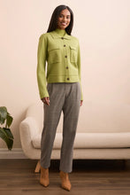 Load image into Gallery viewer, Woman wearing a green jacket, gray pants and brown boots standing in a room with a beige sofa and plant.
