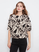 Load image into Gallery viewer, A woman modeling a short-sleeve knit top with a black, brown and 0ff-white cow-print pattern, paired with black pants, against a plain light gray background.