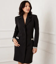 Load image into Gallery viewer, Woman wearing a black coat dress with rhinestone trim on pockets and wrists against a white wall.
