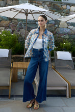 Load image into Gallery viewer, Women wearing blue jeans, gold sandals and floral lace blouse over a white top standing outside with rock wall and patio furniture in the background.