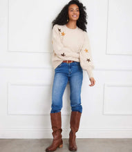 Load image into Gallery viewer, Woman wearing a cream sweater with embroidered flowers, brown knee-high boots and blue jeans against a white wall.