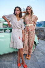 Load image into Gallery viewer, A breathtaking blend of orange, brown and red floral motifs on a pastel peach backdrop define the fashionable aura of the Tinslee jumpsuit. Delicate gold accents adorn the piece, accentuating the visual appeal of the design. For added drama, it features ruffled sleeves and an elasticized waist with tassels to give it a captivating, effortless style. Color- Orange, brown, red, pastel peach, gold. One piece. Elasticized waist with tassels. V-neck. Ruffle sleeves. Fabric- 100% Polyester.