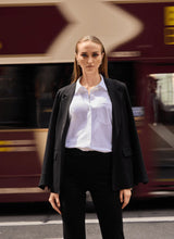 Load image into Gallery viewer, Woman in a black blazer, black pants and white shirt standing in front of a blurred train.
