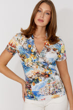 Load image into Gallery viewer, Woman wearing a colorful short-sleeve shirt with a scenic print and white pants on a plain background.