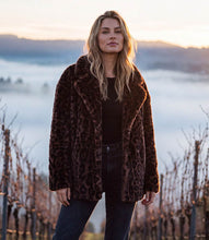 Load image into Gallery viewer, A woman wearing a brown leopard-print faux fur coat over a black top and dark jeans, standing outdoors in a misty vineyard landscape at sunrise or sunset.
