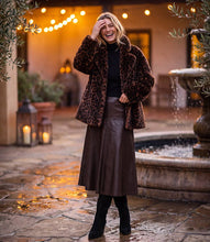 Load image into Gallery viewer, A woman stands in a warm, outdoor courtyard wearing a brown leopard-print faux fur coat over a black top and a brown leather skirt, with black boots. A stone fountain and string lights are blurred in the background.