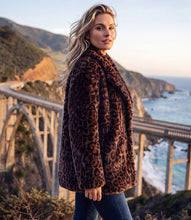 Load image into Gallery viewer, A woman wearing a brown leopard-print faux fur coat and blue jeans stands on a coastal cliffside path with the Bixby Bridge and ocean in the background.