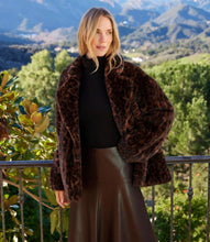 Load image into Gallery viewer, A woman stands on a balcony wearing a dark brown leopard-print faux fur jacket over a black top and brown skirt, with a green mountain landscape in the background.