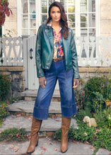 Load image into Gallery viewer, Woman wearing a green leather jacket, colorful blouse, blue jeans, and brown boots standing outdoors.