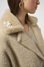 Load image into Gallery viewer, Close-up of a person wearing bold gold earrings and a warm taupe coat with decorative pearl brooch against a plain background.