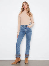 Load image into Gallery viewer, A woman modeling a beige ribbed mock-neck sweater and blue jeans with decorative embroidered cuffs, paired with tan ankle boots, standing against a white wall.