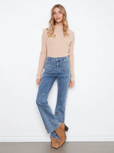 Load image into Gallery viewer, A woman stands against a white wall wearing a beige ribbed mock-neck long-sleeve top, blue high-waisted flare jeans with small embroidered details, and tan ankle boots.