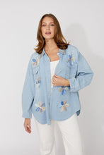 Load image into Gallery viewer, Woman wearing a light blue denim shirt with floral embroidery over a white top and white pants on a white background.