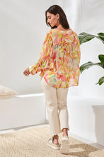 Load image into Gallery viewer, Woman wearing a colorful blouse, white pants and white sandals standing in a bright room with a plant.