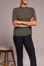 Load image into Gallery viewer, Woman wearing a spruce green short sleeve top, black pants and black boots standing by a barstool against a neutral background.
