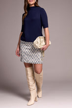 Load image into Gallery viewer, Woman wearing a navy top, patterned skirt, and beige knee-high boots with a beige clutch against a neutral background.