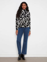 Load image into Gallery viewer, A woman modeling a black and white abstract patterned mock-neck long-sleeve top with blue jeans and black boots, standing against a white wall.
