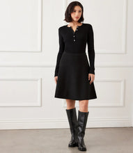 Load image into Gallery viewer, Woman wearing a black ribbed sweater with rhinestone buttons, black skirt and tall black boots against a white wall.