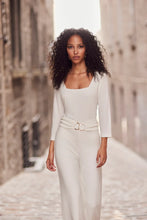 Load image into Gallery viewer, Woman in a white outfit standing in a sunlit alleyway.