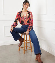 Load image into Gallery viewer, Woman sitting on a wooden stool wearing a colorful long sleeve blouse, blue jeans, and brown boots against a white wall.