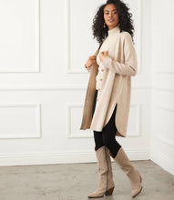 Load image into Gallery viewer, Woman wearing a long beige cardigan over a beige turtleneck, black pants, and tall beige boots against a white wall.