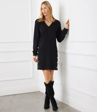 Load image into Gallery viewer, Woman wearing a black dress with gold buttons and black boots against a white wall.