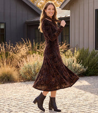 Load image into Gallery viewer, A woman wearing a dark brown velvet long-sleeve midi dress with a subtle floral pattern, paired with black ankle boots, standing outdoors on a cobblestone path in front of a modern house.