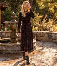 Load image into Gallery viewer, A woman wearing a dark brown floral velvet long-sleeve midi dress and black ankle boots stands on a stone patio in front of a stone fountain, surrounded by trees and warm outdoor lighting.