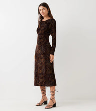Load image into Gallery viewer, A woman modeling a long-sleeve midi dress in dark brown velvet with an intricate floral pattern, paired with black lace-up strappy sandals against a plain light gray studio background.