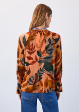 Load image into Gallery viewer, Woman wearing a leaf patterned blouse with long sleeves and gold tie and blue jeans against a neutral background.